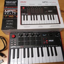 Na fotografii je kompaktní MIDI klaviatura AKAI PROFESSIONAL MPKminiplay v černé barvě s červeným okrajem, umístěná před svým původním kartonovým oba…