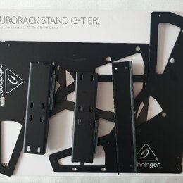 Bílá krabice s nápisem EURORACK STAND (3-TIER) a podtitulem 3-Tier Eurorack Stand for 70, 80 and 104 HP Chassis. Uvnitř jsou rozložené černé kovové d…