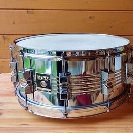 Stříbrný kovový snare drum s chromovaným povrchem. Na boku je černý čtvercový štítek s bílým nápisem MAPEX a menším textem pod ním, který je nečiteln…