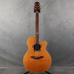 Akustická kytara s vyřezem (cutaway) v přirozené dřevěné barvě. Na hlavě je viditelné logo "Taylor". Hриф má v 12. poličce malý modrý ozdobný prvek.…