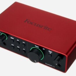 Červený audio rozhraní s kovovým povrchem. Na horní straně je nápis Focusrite a na předním panelu vpravo označení Scarlett 2i2. Zařízení má dva vstup…