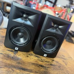 Dva černé studiové reproduktory umístěné na dřevěném stole. Na spodní části obou reproduktorů je bílé logo JBL. Povrch je matný a v horní části se na…