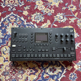 Černý sampler Octatrack MKI s displejem a řadou ovládacích prvků v podobě tlačítek a otočných knoflíků. Na přední straně je bílý nápis Octatrack MKI…