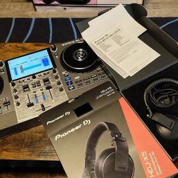 Na fotografii je černý DJ 컨트롤er značky Numark s modrým displejem a dvěma jogovými koly. Vedle něj leží krabice a černé sluchátka s nápisem Pioneer DJ…