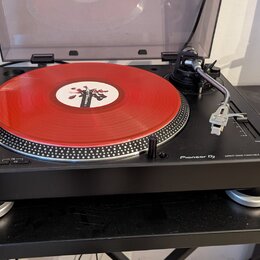 Černý turntable Pioneer DJ s nápisem DIRECT DRIVE TURNTABLE PLX-500. Na talíři je umístěna červená vinylová deska s bílým štítkem. Přístroj je vybave…