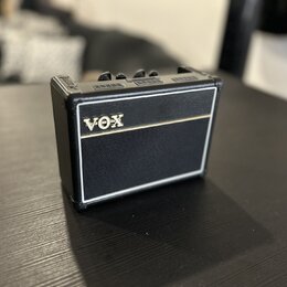 Malý kytarový zesilovač s černým povrchem z texturovaného materiálu a bílými lemovými pruhy. Na přední straně je umístěno zlaté logo VOX. Horní panel…