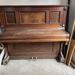 Hnědé dřevěné piano s výraznou masivní konstrukcí a dekorativními panely. Povrch je pokryt prachem a vykazuje známky opotřebení v podobě škrábanců. N…