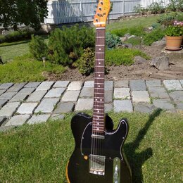 Elektrická kytara s černým bodies, hnědým hnedem na okrajích a tmavým hmatníkem. Na hlavě je viditelné logo Fender Telecaster. Kytara má černý pickgu…