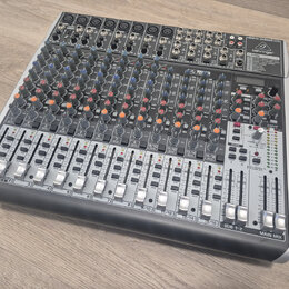 Stříbrný mixážní pult značky behringer s nápisem XENYX QX2222USB. Zařízení obsahuje numerousto potenciometrů v modré a oranžové barvě, posuvníky fade…