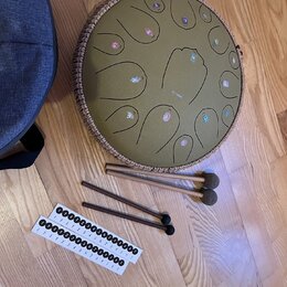Kruhlový steel tongue drum v olivové barvě s hnědým pleteným okrajem. Na povrchu jsou vyryté tóny s barevnými značkami a uprostřed je malý nápis "the…