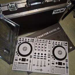 Bílý DJ kontrolér s nápisem TRAKTOR KONTROL S4. Vedle něj je položen hnědý karton s nápisem TRAKTOR KONTROL S4 a v pozadí černý transportní kufr s lo…