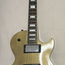 Zlatá elektrická kytara s dvěma humbuckery a čtyřmi ovládacími knoflíky. Na hlavě nástroje není žádný viditelný nápis ani logo. Povrch těla vykazuje…