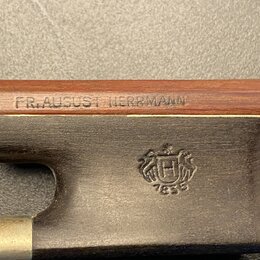 Detail dřevěného těla nástroje s nápisem FR. AUGUST HERRMANN. Pod nápisem je tmavý kovový díl s vyrytou heraldickou značkou, ve které je písmeno H a…