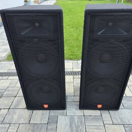 Dvě černé reproduktorové bedny s textilním potahem a kovovými mřížkami. Každý reproduktor má v dolní části oranžové logo JBL. Povrch textilu vykazuje…