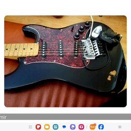 Černá elektrická kytara s červeným mramorovaným pickguardem a světlým hnedým hmatníkem. Nástroj má tři snímače typu single-coil, třetí knoflík a pěti…