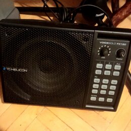 Černý kompaktní reproduktor s kovovou mřížkou. Na přední straně je nápis VOICESOLO FX150 a logo TC-HELICON. Ovládací panel obsahuje dva otočné knoflí…