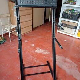 Černý kovový stojan na klávesové nástroje s perforovanou horní deskou. Stojan má nastavitelné výškové mechanismy a dvě boční opěrky pro zajištění nás…