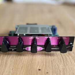 Černý kytarový zesilovač s fialovým předním panelem. Na panelu jsou čtyři černé otočné knofíky s nápisy DRIVE, BASS, MIDDLE PULL/CHANNEL a TREBLE, dá…