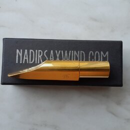 Zlatý kovový mouthpiece na saxofon položený na černé krabičce. Na krabičce je bílý nápis NADIRSAXWIND.COM a na mouthpieceu je vyryté logo TD. Povrch…