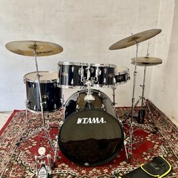Černá bicí souprava s kovovým kováním, umístěná na červeném orientálním koberci. Na basovém bubnu je bílý nápis TAMA®. Souprava obsahuje tři tommy, s…