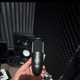 Černý dynamický mikrofon s mřížkou v horní části. Na těle je viditelné logo AKG a pod ním nečitelný nápis modelu. Mikrofon drží ruka, v pozadí jsou v…