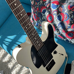 Bílá elektrická kytara typu Telecaster s černým pickguardem a hnědým hmatníkem. Nástroj má dva snímače, jeden ovládací knoflík a přepínač. Na kytáře…
