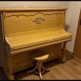 Světle hnědé dřevěné piano s ozdobným vyřezáváním v horní části. Před nástrojem stojí jednoduchá dřevěná taburetka na čtyřnohém podstavci. Povrch pia…