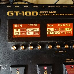 Černý efektový procesor s nápisem GT-100 COSM AMP EFFECTS PROCESSOR. Zařízení má dvě oranžové displeje s texty U01-4, Solo a dalšími zkratkami efektů…