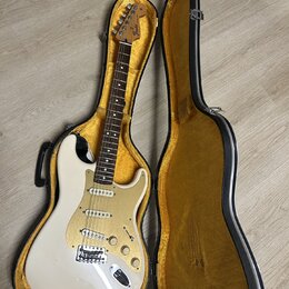 Bílá elektrická kytara s tvarem Stratocaster v černém kufru s žlutým vnitřkem. Na hlavě hředu je viditelné logo Fender. Kytara má tři snímače, zlatý…