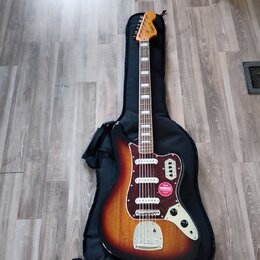 Elektrická kytara s tělem v barvě sunburst a bílým hmatníkem, položená na černém softcasu. Na hlavě je viditelné logo Squier a na těle se nachází čer…