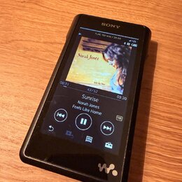 Černý digitální přehrávač značky SONY s displejem. Na obrazovce je zobrazen album cover Norah Jones, název skladby Sunrise a album Feels Like Home. V…
