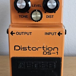 Oranžový kytarový efekt s nápisy Distortion a DS-1. Na horní části jsou tři ovládací knoflíky s popisky TONE, LEVEL a DIST a kontrolka CHECK. Vprostř…