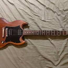 Elektrická kytara s hnědým dřevěným povrchem a černým pickguardem. Na hlavě je viditelné logo Gibson. Kytara má dva humbuckery, čtyři ovládací knoflí…