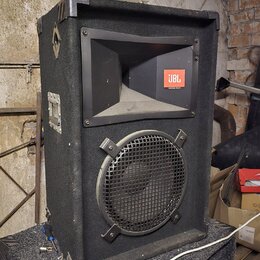 Černý reproduktor v bedně potažené tmavým kobercem s kovovými hranami. V horní části je rohové hrdlo s oranžovým logem JBL, ve spodní části je kruhov…