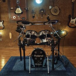 Elektronická bicí souprava značky Roland umístěná na modrém koberci. Na basovém bubnu je bílý nápis Roland a níže logo V-Drums. Souprava obsahuje dva…