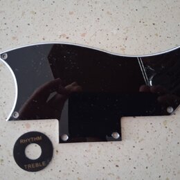 Černý plastový plech ochranný (pickguard) s vyleštěným stříbrným okrajem a otvory pro šrouby. Vedle něj je umístěn malý černý kruhový přepínač s bílý…
