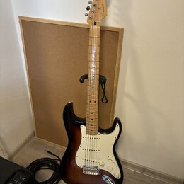 Elektrická kytara s tłem v barvě sunburst a bílým pickguardem. Na hlavě hředu je viditelné logo Fender. Kytara stojí na černém stojanu před korkovou…