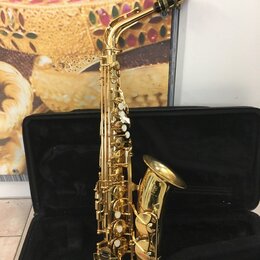 Zlatý saxofon umístěný v otevřeném černém kufru. Na zvonu nástroje je viditelné logo a nápis "YAMAHA". Povrch instrumentu je lesklý, s bílými odpoutá…