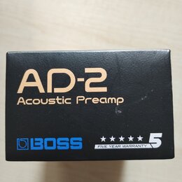 Černá krabice s produktem Boss AD-2. Na přední straně je zlatým písmem nápis AD-2 Acoustic Preamp a modré logo BOSS. V pravém dolním rohu je bílý náp…