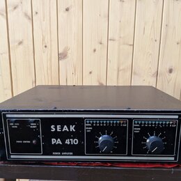 Černý výkonový zesilovač v kovovém šasi s úchopy po stranách. Na předním panelu je bílý nápis SEAK PA 410 a POWER AMPLIFIER. Jsou zde dva ovratidla V…