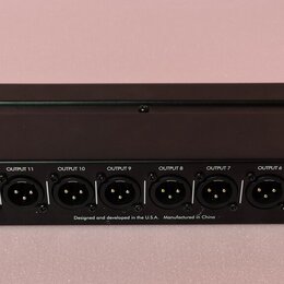 Černý rackový rozdělovač signálu s 16 výstupy typu XLR. Nad každým konektorem je bílý nápis OUTPUT 1 až OUTPUT 16. Na spodní hraně jsou nápisy Applie…