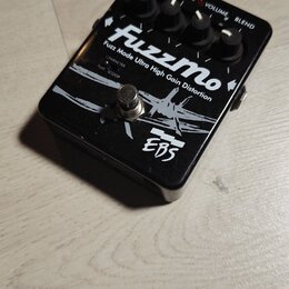 Černý kytarový efekt s bílým nápisem FuzzMo a podtitulem Fuzz Mode Ultra High Gain Distortion. Na spodní části je logo EBS. Zařízení má čtyři ovládac…