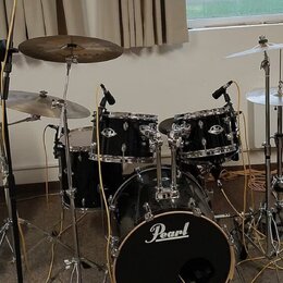 Černá bicí souprava značky Pearl s kovovým hardwarem. Sestává z basového bubnu, dvou tomů a jednoho floortomu, doplněných o tři činely. Na basovém bu…