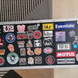 Černý transportní kufr s hliníkovými hranami a kovovými rohy, který je polepen mnoha nálepkami. Mezi viditelnými nápisy a logy jsou: Apple, U2, RADIO…