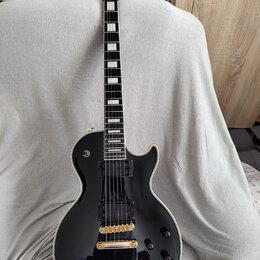 Černá elektrická kytara s tvarem singlecut a bílým lemováním hran. Na hlavě je nápis Epiphone, hриф má vložky z perlece a kytara je vybavena dvěma če…