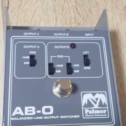 Šedý kovový přístroj s nápisem AB-O BALANCED LINE OUTPUT SWITCHER a logem Palmer Musical Instruments. Na horní straně jsou tři přepínače s popisky PA…