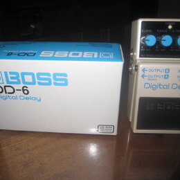 Bílý efekt pedál Digital Delay DD-6 značky BOSS s modrými ovládacími knoflíky. Vedle pedálu je umístěna jeho původní bílá krabice s modrým nápisem BO…