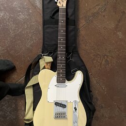 Krémová elektrická kytara typu Telecaster s bílým pickguardem, položená na černém softcasu. Na hlavě hředu je viditelné logo Fender. Kytara má žlutý…