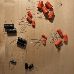 Na dřevěném povrchu je rozprostřeno několik elektronických součástek, konkrétně kondenzátorů. Většina z nich má oranžový povrch a kovové vodiče, osta…