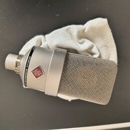 Stříbrný kondenzátorový mikrofon s kovovou síťkovou hlavou, položený na bílé látce. Na těle je vidět logo v červeném kosočtverci a nápis NEUMANN MICR…